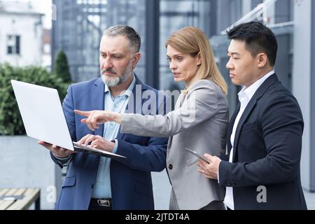 Successful business team, three colleagues businessman and businesswoman outside office building discussing current plans and management, looking at laptop screen, discussing and consulting Stock Photo