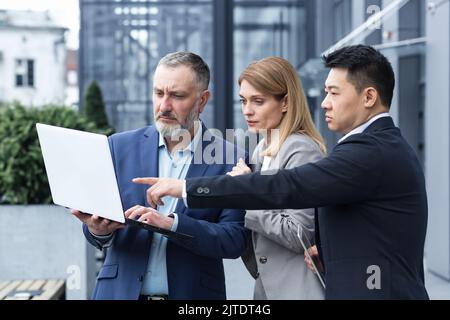 Successful business team, three colleagues businessman and businesswoman outside office building discussing current plans and management, looking at laptop screen, discussing and consulting Stock Photo