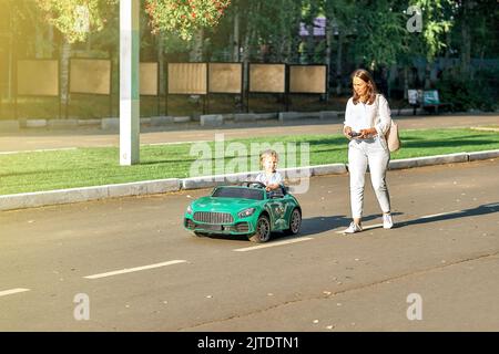 Mom controls children car with remote at walk with toddler child in public park. Toddler boy sits in green car holding steering wheel looking aside Stock Photo