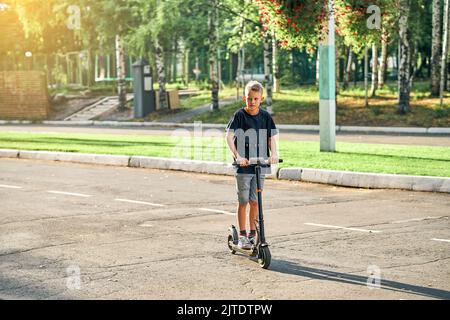 Schoolboy rides electric scooter on road in empty summer park. Child in black t-shirt and denim shorts rides on scooter along marked asphalt road Stock Photo