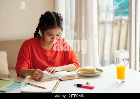 Confused schoolgirl having sandwich and glass of juice when reading book for English class Stock Photo