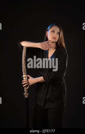 Vertical portrait of a pensive young brunette woman with a katana on a black background Stock Photo