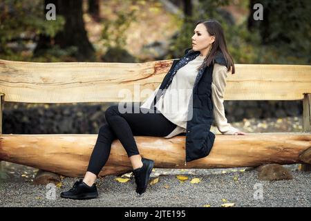 Profile of a young woman in full growth sitting on a wooden bench in a park outdoors Stock Photo