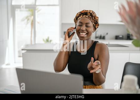Beautiful young Black African woman wearing tradition headscarf. Sitting at home working on laptop. On phone call smiling and gesturing with hand Stock Photo