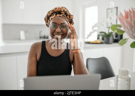 Black African woman smiling, holding eye glasses. Looking at laptop while sitting and working from home. Wearing traditional head scarf Stock Photo