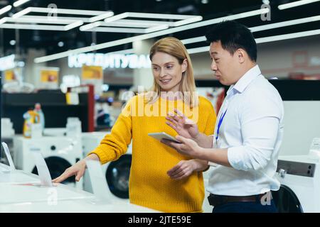 Asian consultant salesman in electronics and household appliances store, selling a working machine to a woman, recommending and approving the choice Stock Photo