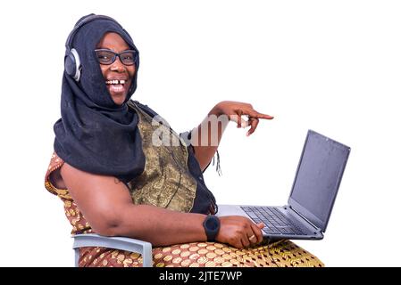 beautiful muslim businesswoman sitting on white background with headset pointing at laptop and looking at camera laughing. Stock Photo