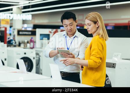 Asian consultant salesman in electronics and household appliances store, selling a working machine to a woman, recommending and approving the choice Stock Photo