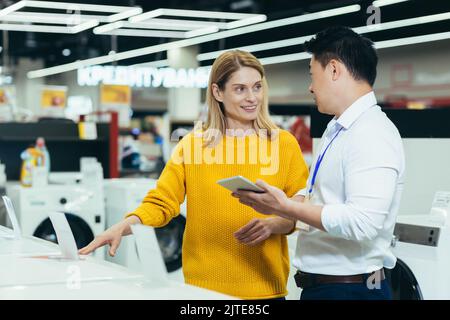 Asian consultant salesman in electronics and household appliances store, selling a working machine to a woman, recommending and approving the choice Stock Photo