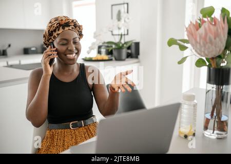 Beautiful young Black African woman wearing tradition headscarf. Sitting at home working on laptop. On phone call smiling and gesturing with hand Stock Photo