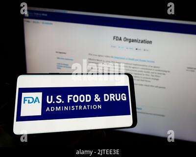 Person holding mobile phone with logo of United States Food and Drug Administration (FDA) on screen in front of web page. Focus on phone display. Stock Photo