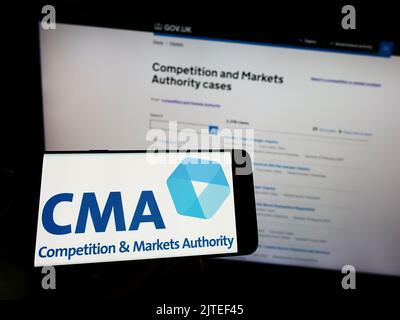 Person holding mobile phone with logo of British Competition and Markets Authority (CMA) on screen in front of web page. Focus on phone display. Stock Photo