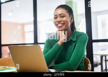 Happy inspired African-American female office employee using laptop indoors, resting chin with hand and looking at camera, smiles. Excited freelancer woman enjoys remote work in coworking space Stock Photo