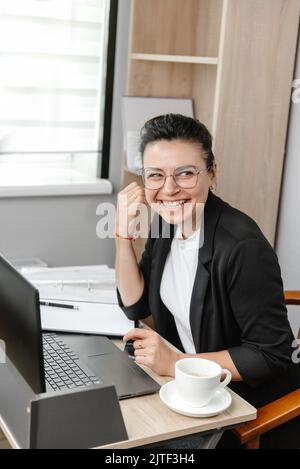 A young businesswoman employee or manager uses a computer, looks at a customer or employee and smiles. Hybrid work empty office Stock Photo