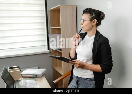 A girl works in an office. The office worker is pensive and looks away. Hybrid job Stock Photo