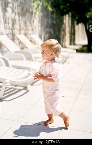 Little girl in summer overalls stands on a tile near white sun loungers Stock Photo