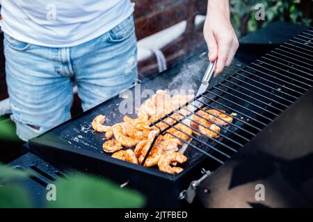 Grilling shrimp on skewer on outdoor grill. Grilled shrimps on the flaming grill. Man hand using tong pinching grilled seafood Stock Photo