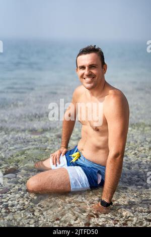 Young smiling man sitting in the water near the shore Stock Photo