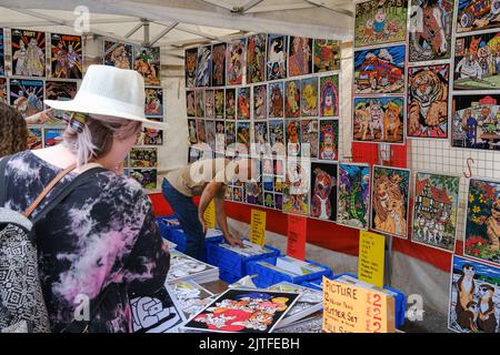 Ballycastle, United Kingdom. 30 Aug, 2022. Woman in white hat and summer dress browsing art prints on stall at the closing day of Ireland's oldest fair in the seaside town of Ballycastle on the north coast of North Antrim wrapping up a bank holiday weekend of trading, and a programme packed with entertainment, local and international cuisine at The Ould Lammas Fair. Credit: Steve Nimmons/Alamy Live News Stock Photo