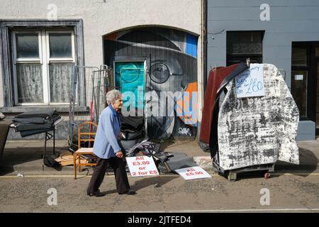 Ballycastle, United Kingdom. 30 Aug, 2022. Woman walks past mural and stack of carpets for sale in street. The closing day of Ireland's oldest fair in the seaside town of Ballycastle on the north coast of North Antrim wrapping up a bank holiday weekend of trading, and a programme packed with entertainment, local and international cuisine at The Ould Lammas Fair. Credit: Steve Nimmons/Alamy Live News Stock Photo