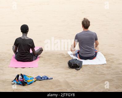Mixed race couple performing yoga, meditation on beach. Stock Photo