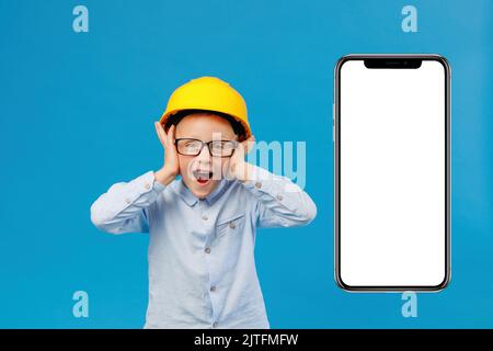 A cute boy construction worker with a shocked expression in a yellow hard hat is standing in an indoor studio on a blue background near huge mobile ph Stock Photo