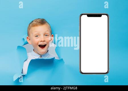 Happy cute boy is having fun played on blue background wall, climbs through a hole in the paper near huge mobile phone. Bright and funny emotions of t Stock Photo
