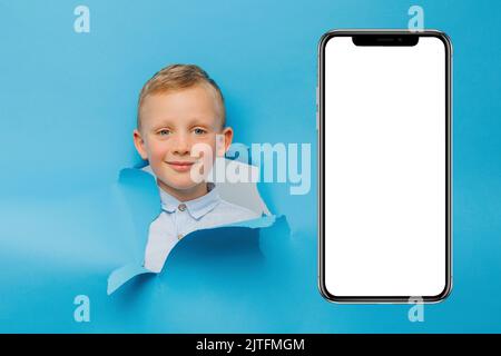 Happy cute boy is having fun played on blue background wall, climbs through a hole in the paper near huge mobile phone. Bright and funny emotions of t Stock Photo