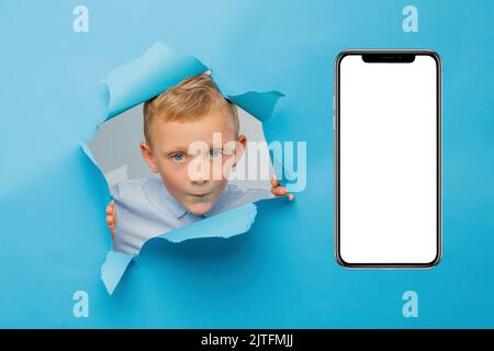 Happy cute boy is having fun played on blue background wall, climbs through a hole in the paper near huge mobile phone. Bright and funny emotions of t Stock Photo