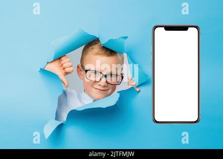 Happy cute businessboy in eyeglasses is having fun on blue background wall, climbs through a hole in the paper near huge mobile phone. Bright and funn Stock Photo