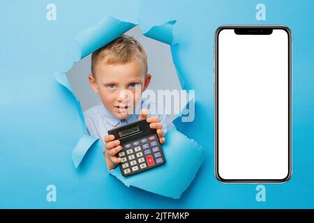 Happy cute businessboy is having fun and holding calculator in hand on blue background wall, climbs through a hole in the paper near huge mobile phone Stock Photo
