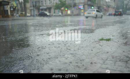 Raindrops fall on the road with cars driving in the background. Heavy torrential rain in the city. Stock Photo