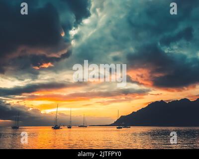 Dramatic nature sunset in tropical island lagoon. Yachts silhouette against bright orange blue clouds in sky. Ocean calm water and mountain hill. Wonderful picturesque scene. Majestic travel landscape Stock Photo