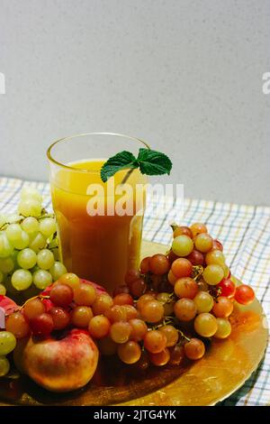 Multifruit juice in a faceted glass stands on a table among the various juicy fresh tropical fruits, grapes in summer. A healthy and refreshing drink. Stock Photo