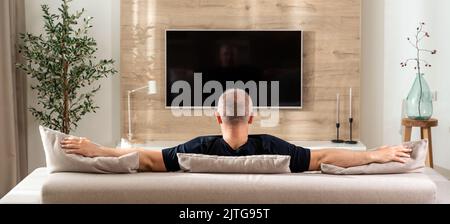 Man spread his arms on the sofa in front of the TV in a modern living room Stock Photo