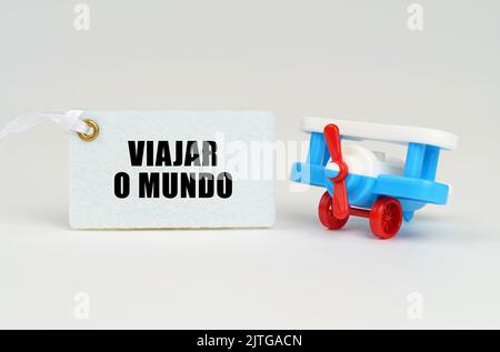 On a white surface there is a toy airplane and a sign with the inscription - Travel the world. Text in Portuguese. Stock Photo