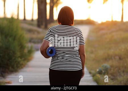 back view of active overweight woman with yoga mat walking to beach to practice yoga, exercising outdoors on sunrise in the morning. Concept of health Stock Photo