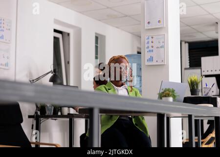 African american employee working, sitting at coworking open space desk, colleagues on background. Young elegant woman typing on computer at workplace table, low angle ciew Stock Photo