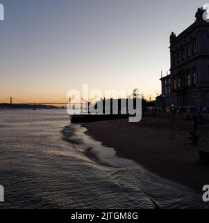 Lisbon beach on the River Tagus as the sun sets on a summers evening with the Ponte 25 de Abril bridge behind. Stock Photo
