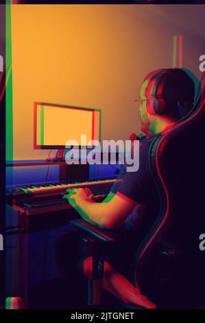 rear view vertical image of bearded young caucasian man, wearing glasses and headphones, sitting working on his home music recording set, typing on co Stock Photo