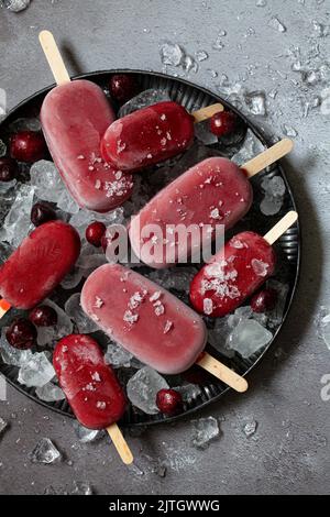 Large and miniature healthy cherry popsicles (ice pops) on a plate of ice with frozen cherries scattered on top. Stock Photo