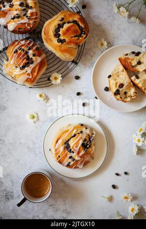 English currant buns, some glazed with icing and some unglazed. One is on a plate with an espresso coffee beside it. Stock Photo