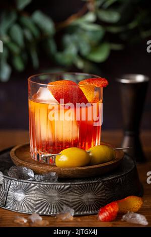 A Negroni cocktail made with orange coloured italian Bitters and garnished with orange zest. Two green olives on a cocktail pick rest at the side.. Stock Photo