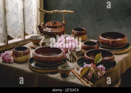 Japanese Asian style table setting with empty craft ceramic tableware, brown rough bowls, craft teapot and cups on linen tablecloth, decorated by pink Stock Photo