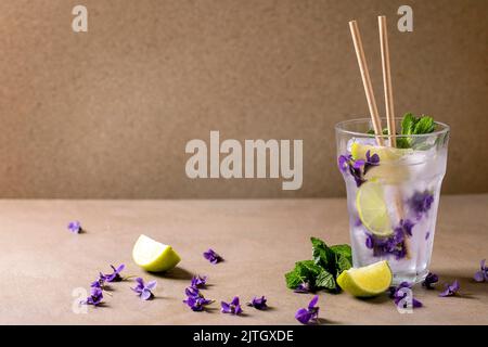 Transparent glass of lemonade soda cocktail with mint, violets flower ice and lime slices, paper straws, standing on brown table. Cold summer drink Stock Photo