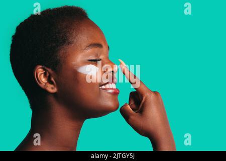 Profile view of african woman touching nose with index finger to apply cream isolated on sea green background with free space for text. Facial mask Stock Photo