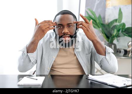 Stunned puzzled african american man with headset, business leader or coach, talking with colleagues by video call, discuss ideas and promotion, looks at camera in confusion, gesturing hands Stock Photo