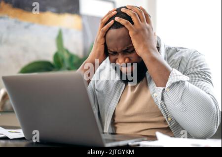 Frustrated sad stressed african american man, sit at desk, uses laptop for online work, annoyed with bad result of project, failed the deal, experiences negative emotions, stress,holding hands on head Stock Photo