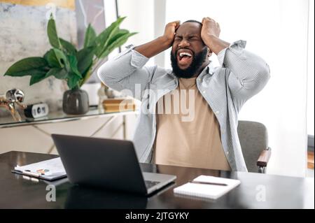 Annoyed african american young man, company worker, freelancer, ceo company, sitting at a desk in modern office, yelling, screaming, dissatisfied with the result, irritated emotional state Stock Photo
