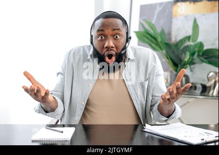 Surprised shocked african american man with headset, business leader or coach, conducts business meeting with colleagues, discuss ideas and promotion, looks at camera in confusion, gesturing hands Stock Photo